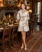 Load image into Gallery viewer, A woman wearing a cream-colored short dress with a brown and gold floral toile pattern stands in a warmly lit dining room with a fireplace, dining table, and chairs.